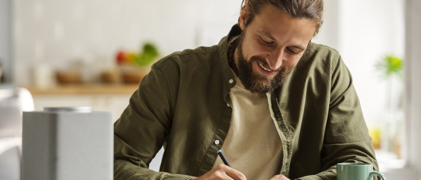 Homen sentado à mesa escrevendo em um caderno, sorrindo, com notebook e caneca ao lado em ambiente doméstico iluminado