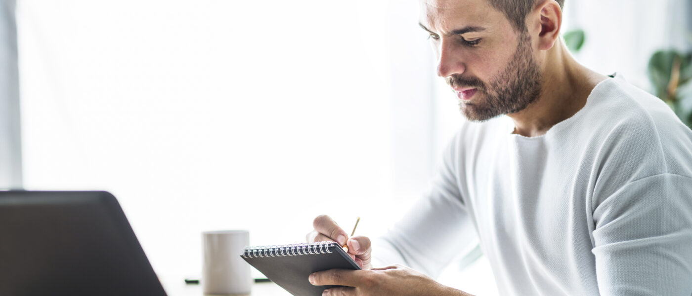 Um homem barbudo em uma camisa branca escrevendo em um pequeno caderno espiral com um lápis, sentado em uma mesa com um laptop e uma xícara.