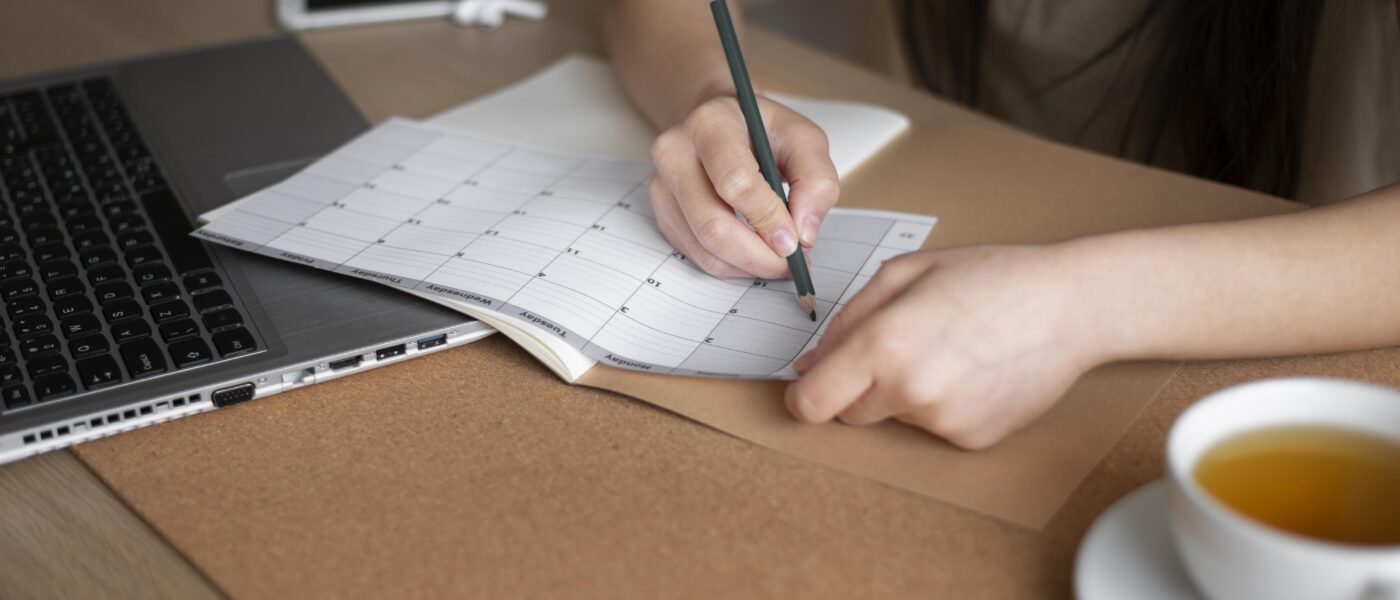 Uma pessoa escrevendo em uma folha de calendário semanal com um lápis, perto de um laptop e uma xícara de chá.
