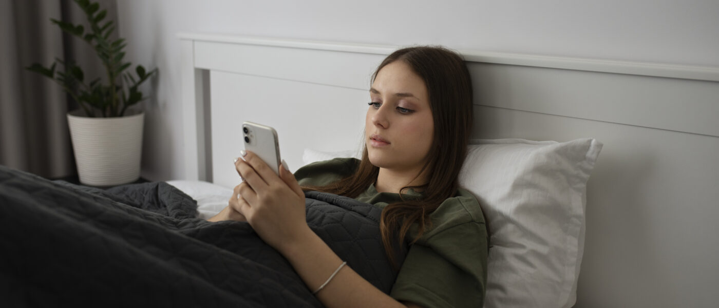 Uma jovem deitada na cama com uma camisa verde, olhando para seu celular.