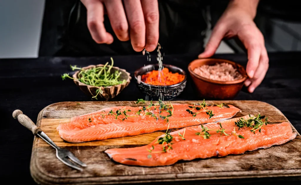 Pessoa temperando um filé de salmão cru com azeite, sal e ervas frescas, preparando o peixe antes do cozimento.