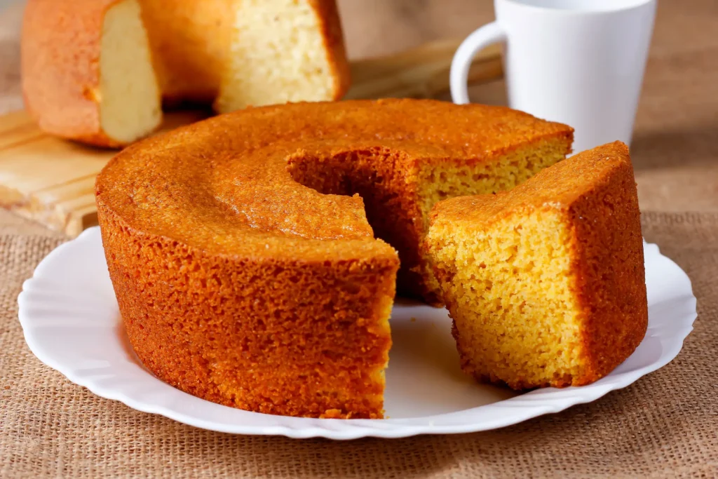 Uma fatia de bolo de fubá é apresentada em destaque, evidenciando sua textura macia e coloração amarela característica.