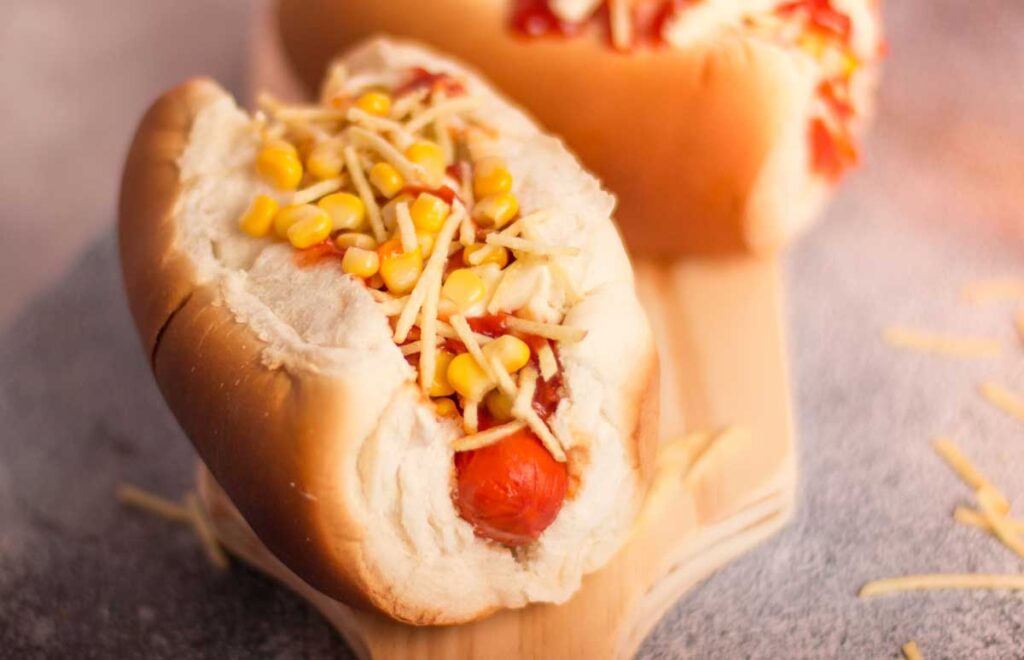Um cachorro-quente é apresentado em destaque, com pão macio envolvendo uma salsicha coberta por milho e batata palha.