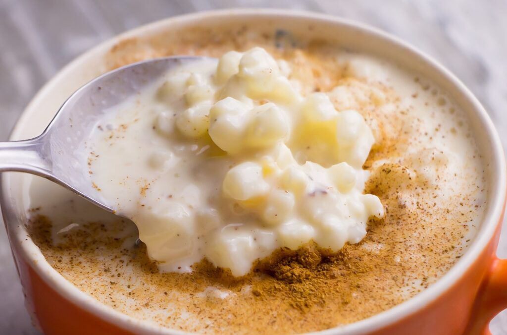 Uma porção de canjica cremosa é mostrada em destaque. Os grãos de milho branco aparecem envolvidos por um creme espesso e brilhante e a superfície é finalizada com canela em pó.