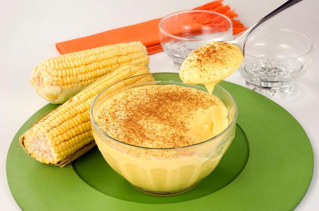 Uma tigela de curau de milho sobre uma mesa de madeira, com textura cremosa e cor amarelo-clara característica. A superfície está polvilhada com canela em pó.
