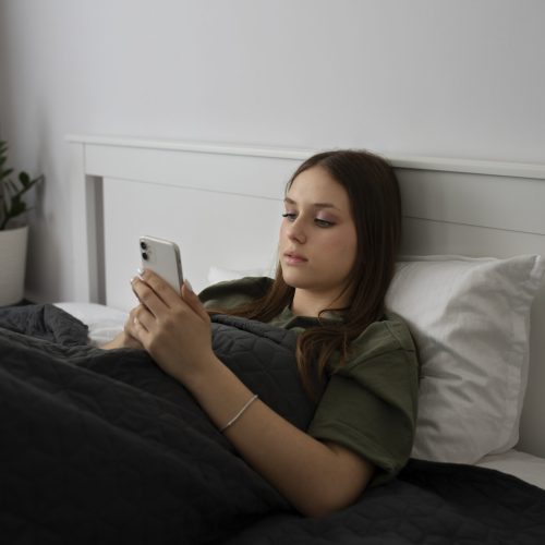 A imagem mostra uma jovem deitada na cama, aparentemente em um quarto. Ela tem cabelos castanhos longos e está vestindo uma camiseta verde-oliva. Seu olhar está fixo no smartphone branco que ela segura com as duas mãos. Ela está coberta por um edredom cinza escuro, com um travesseiro branco visível atrás de sua cabeça. Ao fundo, um vaso de planta verde pode ser visto no canto esquerdo, adicionando um toque de cor natural ao ambiente predominantemente neutro do quarto. A iluminação é suave, sugerindo um ambiente interno tranquilo.