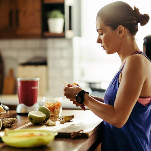 Young sportswoman making herself a healthy smoothie and slicing fresh fruit in the kitchen.